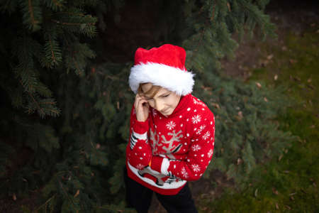 A boy in a red knitted Christmas sweater with a Christmas reindeer and a Santa Claus hat cringed from the cold against the winter skyの写真素材