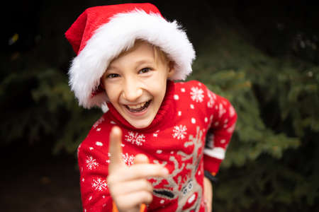 A boy in a red knitted Christmas sweater with a Christmas reindeer and a Santa Claus hat waves his index finger at the camera and laughsの写真素材