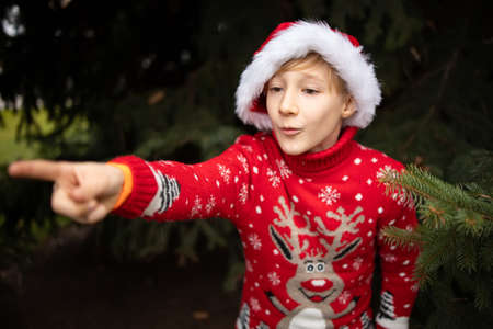 A boy in a red knitted Christmas sweater with a Christmas reindeer and a Santa Claus hat points his finger to the sideの写真素材