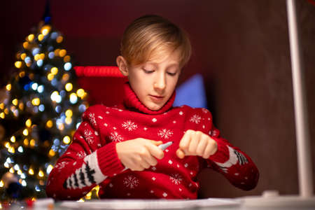 a boy in a red Christmas sweater on the background of a Christmas tree draws in front of a lamp at a white tableの写真素材