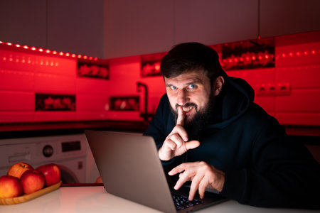 a male hacker behind a laptop in a dark room put his finger to his lips