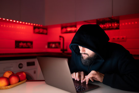 a hacker in a black hoodie with a hood on his head is sitting at a laptop bent over hacking the Internetの写真素材