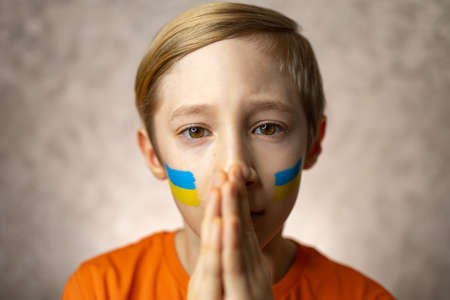 a child against war, a boy with a painted flag of Ukraine on his cheeks folded his hands in a plea for world peaceの写真素材
