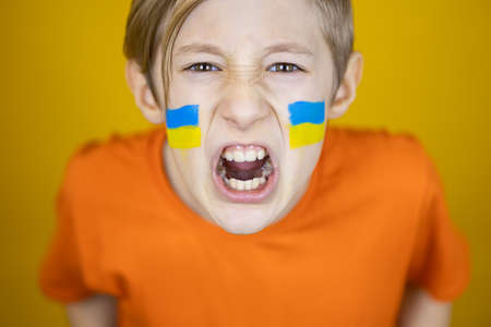 a boy with a painted flag of Ukraine on his cheeks shouts angrily at the camera against the warの写真素材