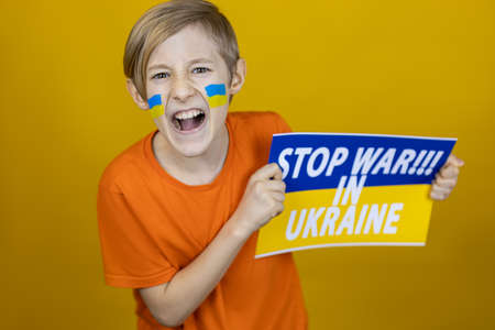 a boy with a painted flag of Ukraine on his cheeks holds a poster on the side with the inscription stop the war in Ukraine and shoutedの写真素材