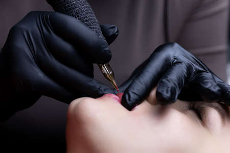 the master performs the procedure of permanent lip makeup in black sterile gloves holds the lips of the model with his fingers stretches them and applies permanent makeup with a tattoo machineの写真素材