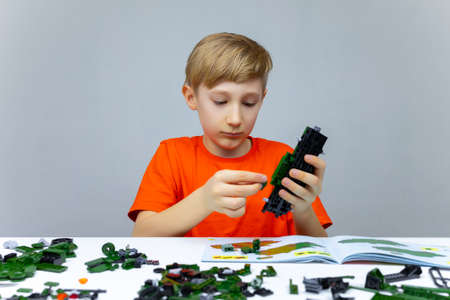 a boy at the table plays with a plastic constructor holds a finished model of the constructor in his...