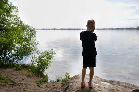 boy traveler stands with his back on a stone and looks pensively at the riverの写真素材