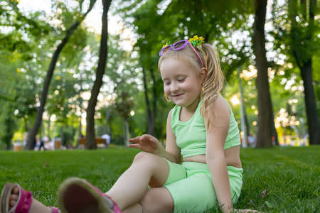 a little girl in a green tracksuit sits on the grass in the park and wipes her kneeの写真素材
