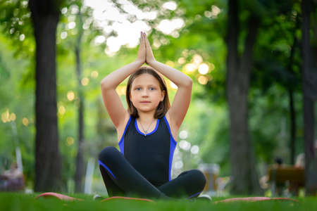 little girl doing yoga sits on the lawn in the park in a twine and looks at the camera