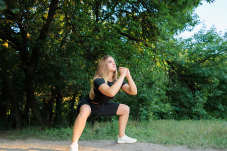slender girl fitness model in a black tracksuit performs squats holding her hands in front of herの写真素材