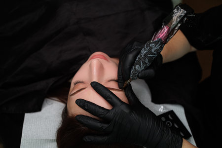 The master has a permanent makeup machine in his hands and he performs the procedure. Eyebrow permanent makeup cosmetic procedure.の写真素材