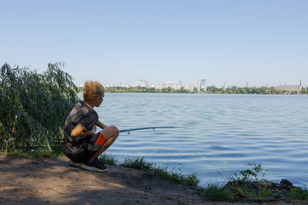 On the bank of a river, a boy is fishing while watching a float. Sport fishing on the river in summer.の写真素材
