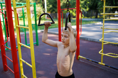 A teenager with an athletic build holds on to the handrails of a sports equipment. Street workout on...