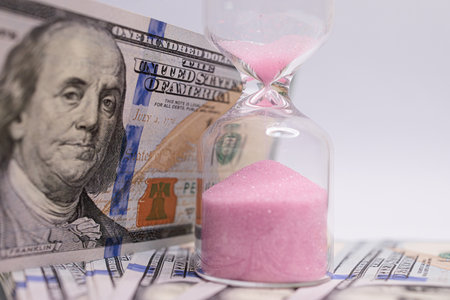 Behind the hourglass is a $100 bill. The hourglass is located near the dollar bills.の写真素材