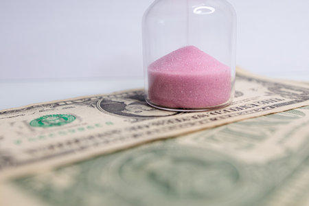 Close-up of sand from the hourglass that stands on dollar bills. The hourglass is located near the dollar bills.の写真素材