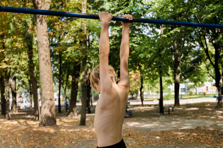 An athletic teenage boy hangs from a crossbar and looks down. Athletic teenage boy in the park...
