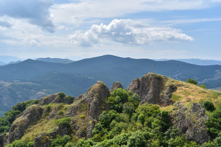 A scenic mountain ridge with a metal cross on top, surrounded by green trees and distant forested hills. Captured under a cloudy sky in a natural landscape. Viewpoint suggests elevation and openness.の写真素材