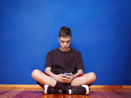 near the blue wall sits a boy and is read on a smartphone. texting message.の写真素材