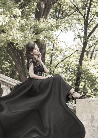 Beautiful girl in a black evening dress sits on the stair railing of an abandoned palace. Beautiful woman with scars. Retro style young girl. Pretty Womanの写真素材