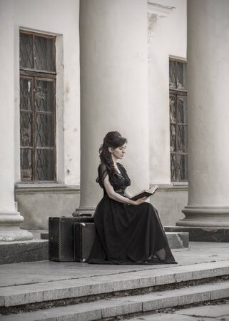 Beautiful girl in black evening sitting reading a book. A girl sits with suitcases near an old building with columns. Beautiful woman with scars. Retro style young girl. Pretty Womanの写真素材