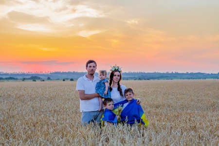 Happy smiling family in national Ukrainian embroidered shirts hold large yellow-blue flag against background of spikelets of wheat field. Patriot education. independence day. Glory to Ukraineの写真素材
