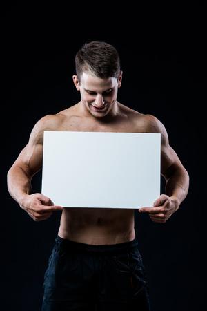 Young strong athlete holding a blank board on dark background looking to the camera and smilingの写真素材