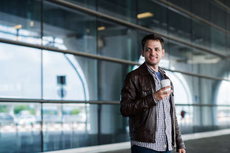 Stylish handsome young male traveller with bristle standing outdoors. Man wearing jacket and shirt. Smiling man looking to camera holding cup of coffee.の写真素材