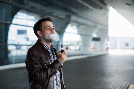Young man enjoying a satisfying e-cigarette standing in profile against airport terminal background.の写真素材