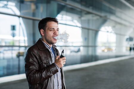 Man with a bristle smoking e-cigarette vaporizer box mode outdoors near the airport terminal before flight.の写真素材