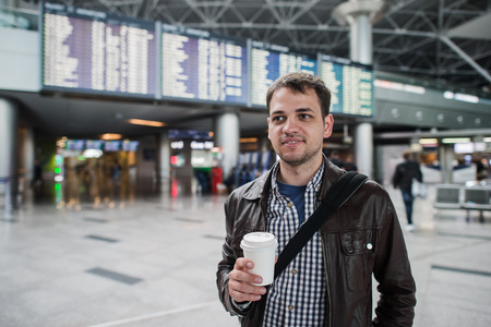 Young man with a bag in airport near flight timetable holding cup of coffee.の写真素材