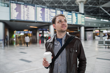 Smiling thoughtful man at board of arrivals and departures at the airport with a cup of coffee.の写真素材