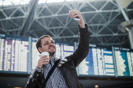 Smiling young traveller man with a coffee at the airport makes selfie in front of timetable board.の写真素材