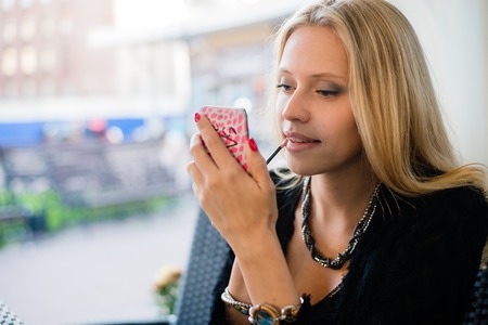 Side portrait of a smart young woman using mirror to apply lipstick cosmetics while visiting a city streetの写真素材