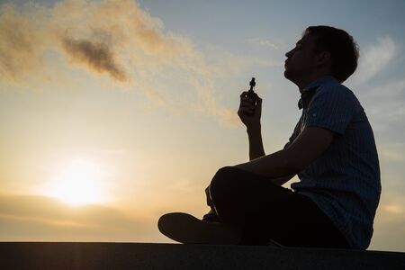 A man smoking vaping e-cigarette vape box mode over golden sunset.の写真素材