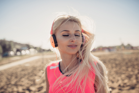 Portrait of young beautiful woman listening to music at beach. Close up face of smiling blonde woman with earphone looking at camera. Girl running at beach and listening to musicの写真素材