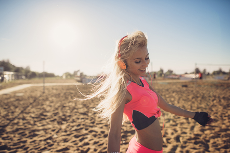 sport outdoor photo of beautiful young blonde woman in pink colorful sport suit listening to music on headphones by the beach.の写真素材