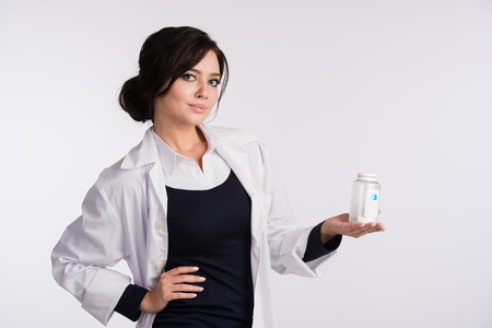 Beautiful young brunette female nurse or physician in blue dress and white lab coat holding a bottle of pills.の写真素材