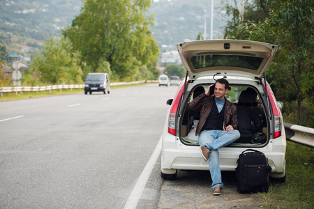 Calling friends. Happy young man talking on the mobile phone and sitting on a car trunk.の写真素材