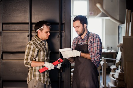Happy male carpenter showing something to coworker ruler and notebook in workshop.