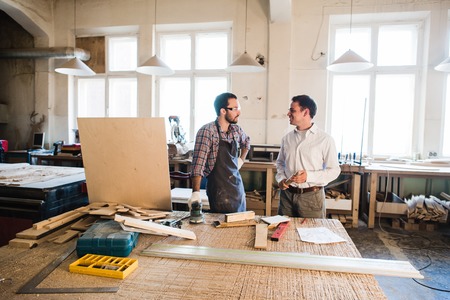 Happy male carpenter showing something to coworker carrying wooden planks in workshop.の写真素材