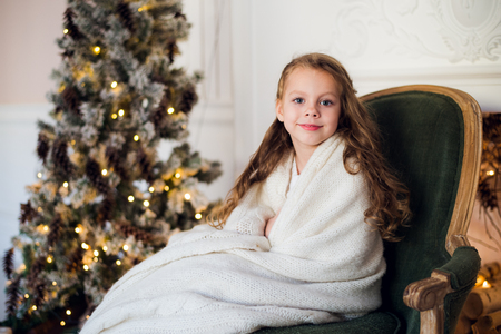 Cute little girl sits by the christmas tree, wrapped in a warm blanket at home.の写真素材