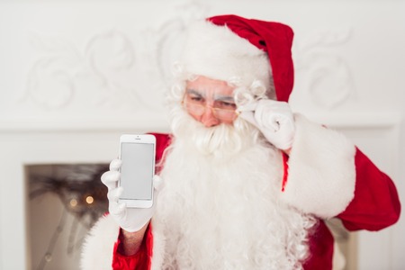 Santa Claus shows a smartphone against red background.の写真素材