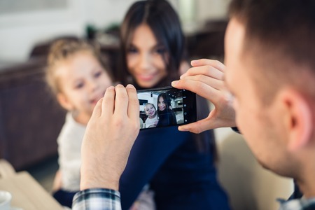 family, parenthood, technology, people concept - happy father taking photo of his little daughter and wife by smartphone having dinner at restaurant.の写真素材