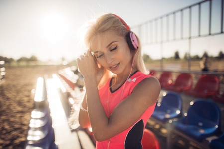 sport outdoor photo of beautiful young blonde woman in pink colorful sport suit listening to music on headphones by the beach.の写真素材