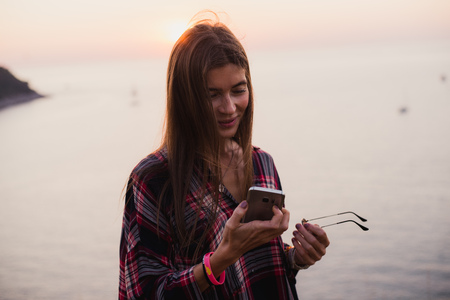 Portrait of young happy woman in hipster shirt standing in front of picturesque sea view, taking selfie with sunset or sunrise on smartphoneの写真素材