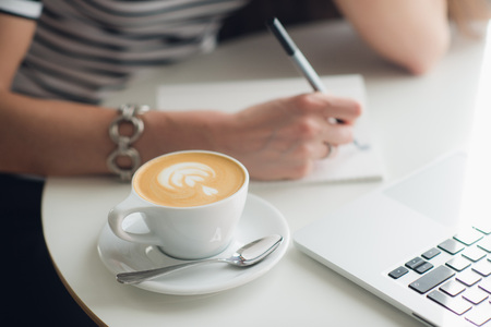 Close up picture of womans hands and a cup of cappuccino. Lady is writing in her notebook with a laptop nearby.の写真素材