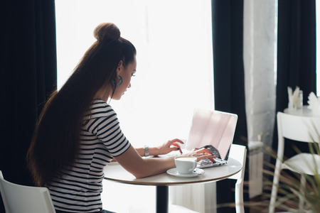 Silhouette of a woman typing on her laptop in front of a window with a cup of coffee.の写真素材