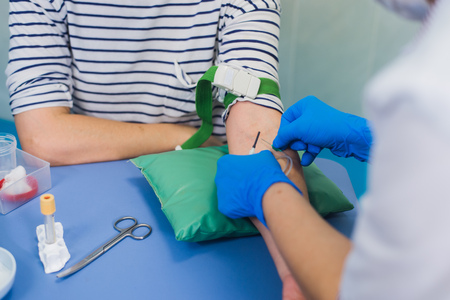 Preparation for blood test with pretty young blond woman by female doctor in white coat medical uniform on the table in white bright room. Nurse pierces the patients arm vein with needle blank tube.の写真素材