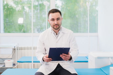 Cheerful young doctor holding a clipboard and gesturing with his hand at hospital ward